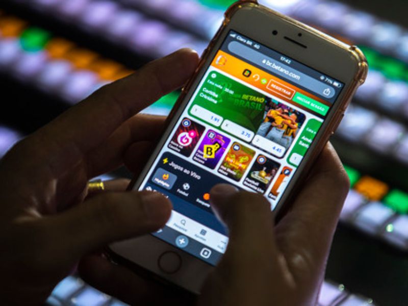 Pessoa utiliza smartphone para acessar aplicativo de apostas esportivas em ambiente com telas iluminadas ao fundo