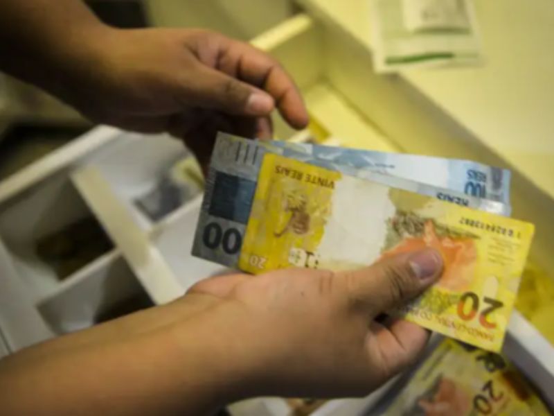 Pessoa contando cédulas de real, com notas de R$ 20, R$ 50 e R$ 100, em ambiente interno.