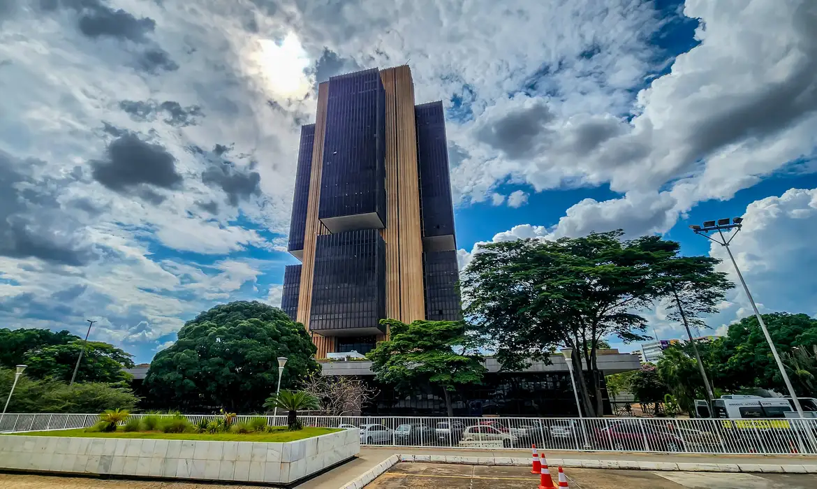 Fachada da sede do Banco Central em Brasília com céu nublado e árvores ao redor.