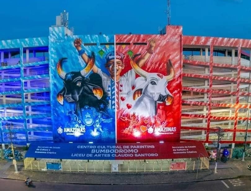 Fachada iluminada do Bumbódromo de Parintins exibe grandes murais dos bois Caprichoso e Garantido, com luzes azul e vermelha representando cada lado da arena.