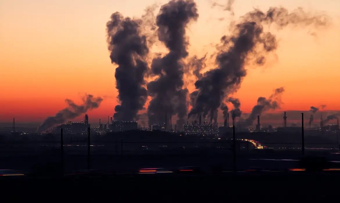 Chaminés industriais liberando grande quantidade de fumaça ao entardecer, com o céu alaranjado ao fundo.