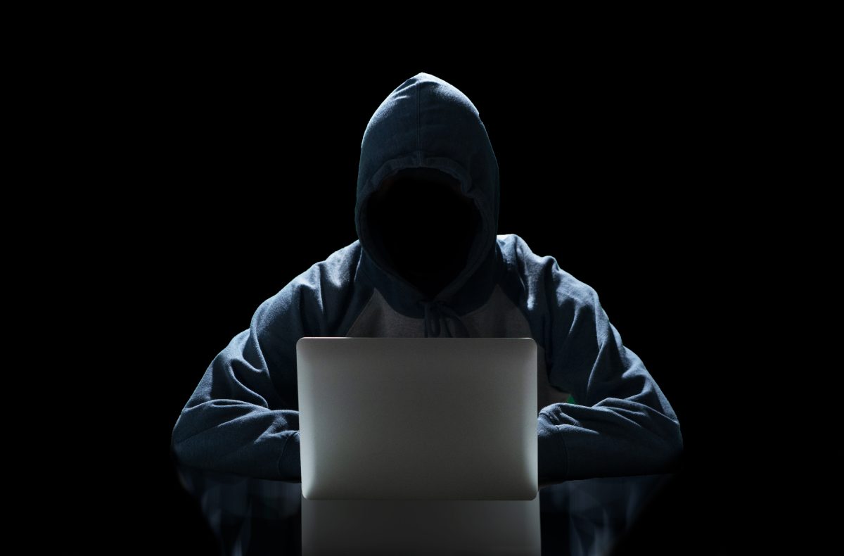 Figura anônima e sombreada de um hacker vestindo um capuz, sentado em frente a um laptop com a tela brilhando, em um cenário escuro, representando a ameaça de ataques cibernéticos