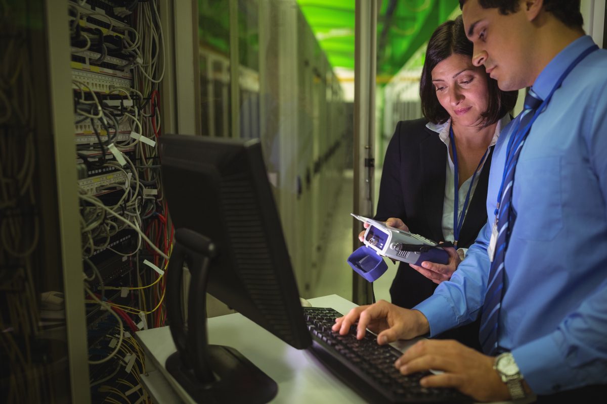 Dois profissionais de TI trabalhando juntos em um computador, com equipamentos eletrônicos e cabos ao fundo, simbolizando a inovação e colaboração no setor.