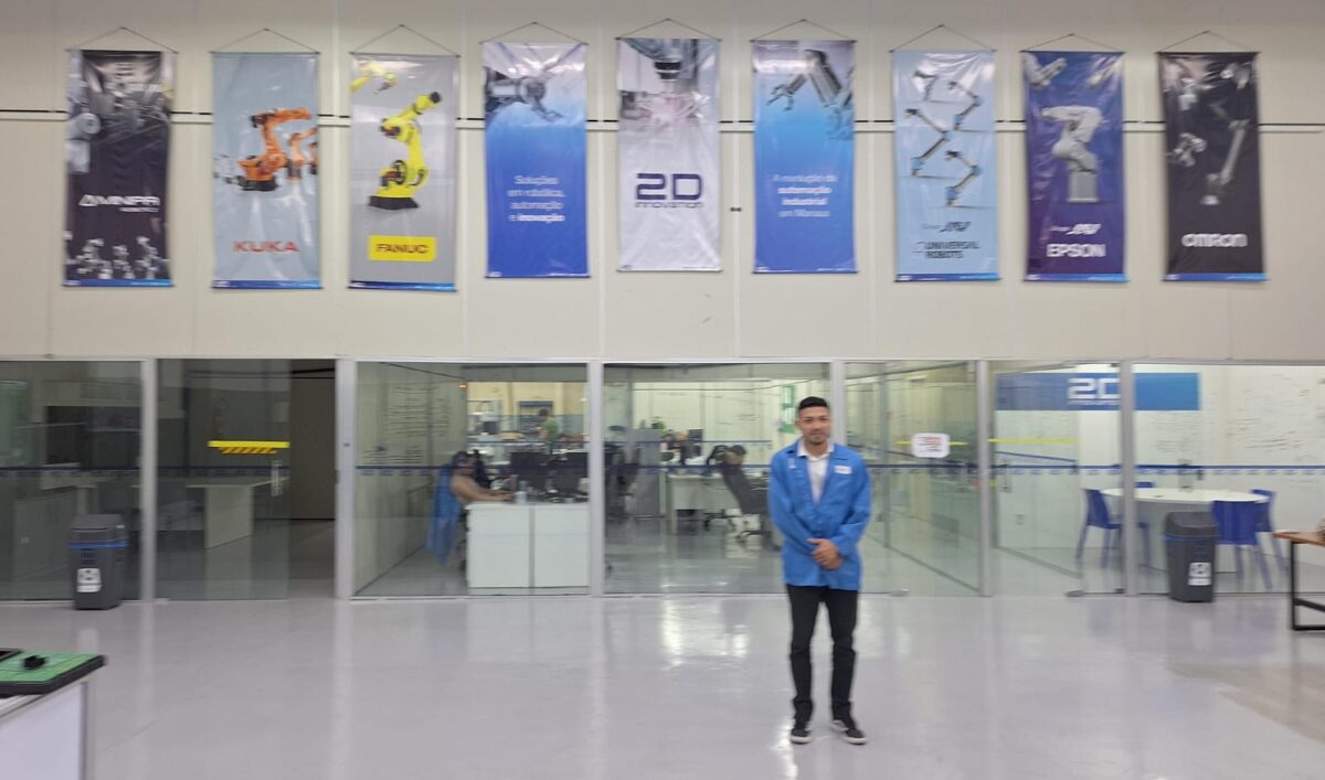David Cunha, fundador da 2D Innovation, posa em frente ao setor administrativo da empresa, com banners de fabricantes de robôs industriais expostos acima.