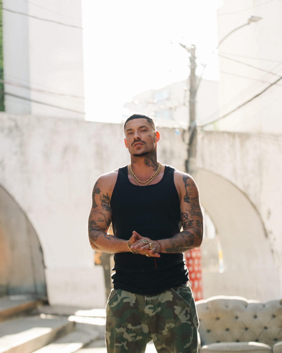 O rapper Filipe Ret posa ao ar livre, vestindo regata preta e calça camuflada, com expressão séria e tatuagens à mostra.