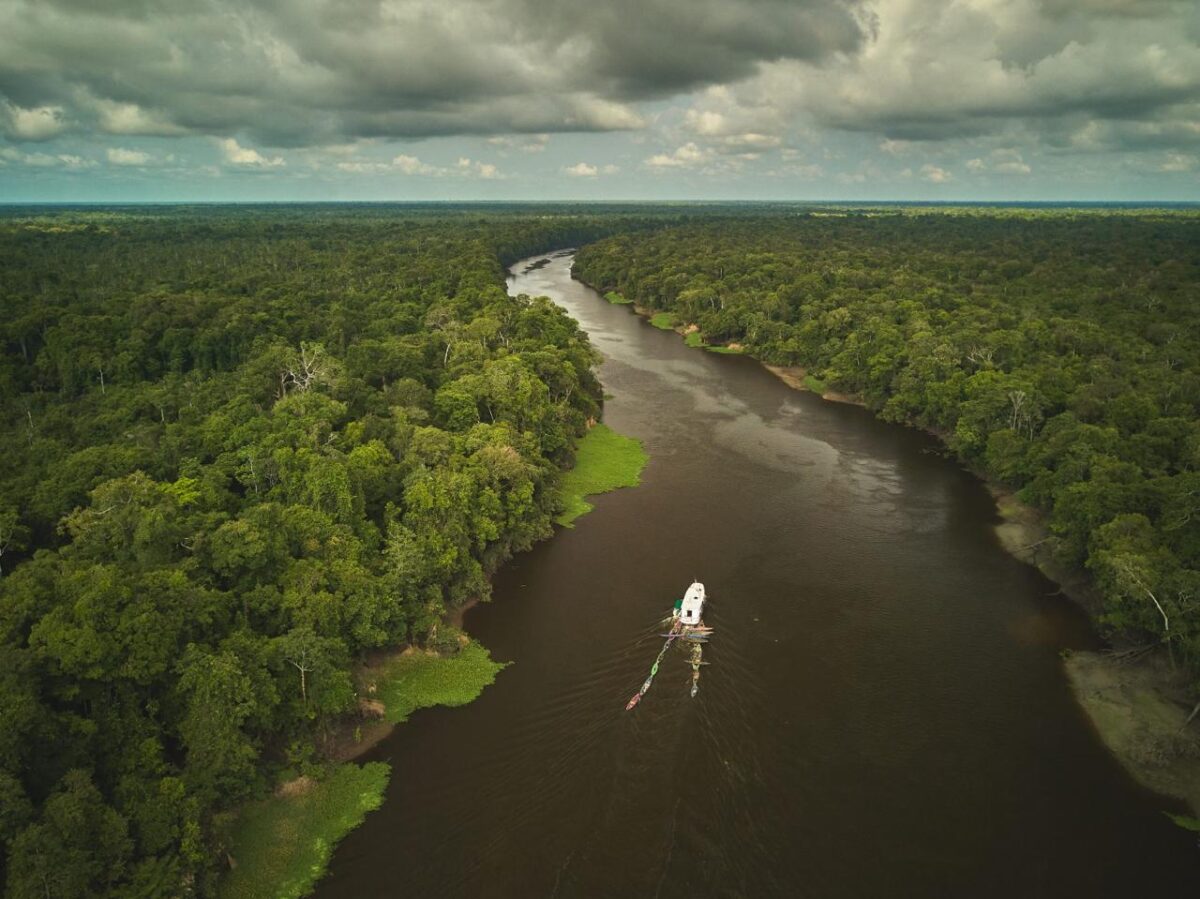 Vista aérea de um barco navegando por um rio sinuoso cercado por floresta densa na Amazônia sob céu nublado.