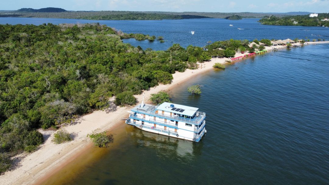 Vista aérea de um barco-laboratório atracado em uma praia amazônica cercada por vegetação e rios, durante preparação da Caravana Iaraçu rumo à COP30 em Belém.