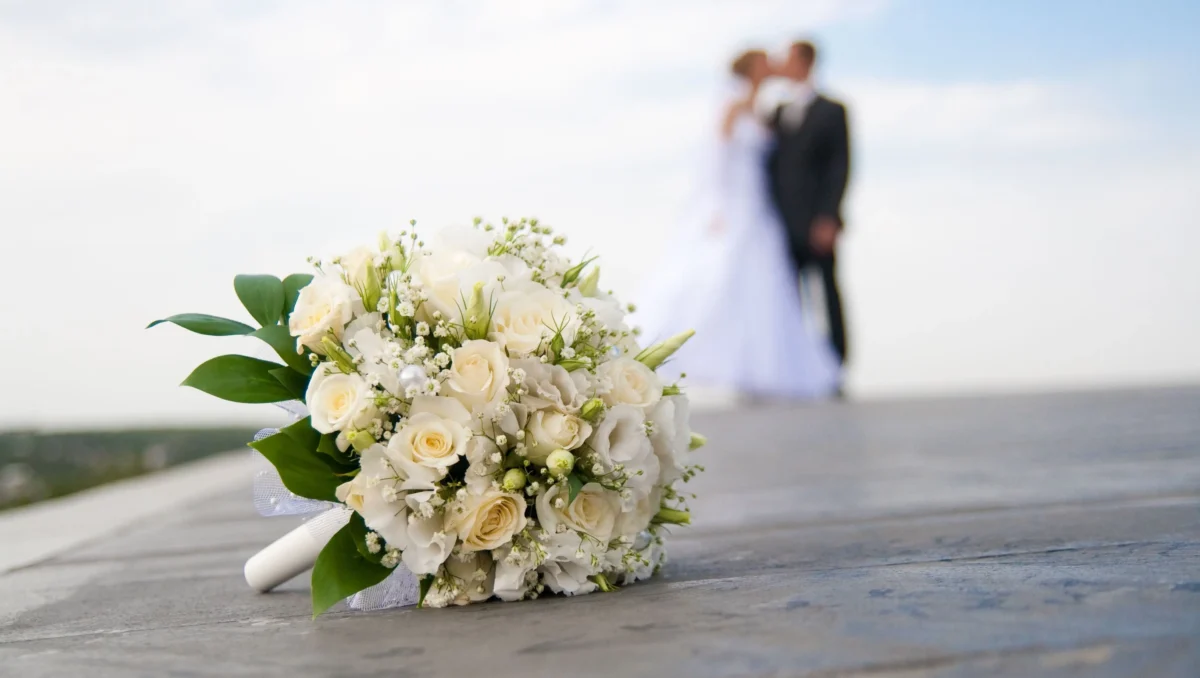 Buquê de flores em primeiro plano com casal de noivos ao fundo durante cerimônia de casamento.