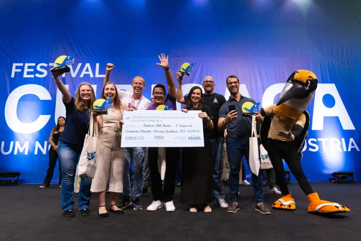Grupo de vencedores do Festival Curicaca 2025 celebra no palco com troféus e cheque simbólico de R$ 15 mil, ao lado do mascote da competição.