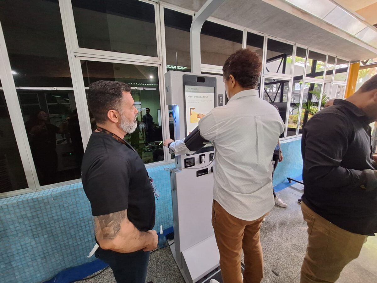 Homem realiza medição de pressão arterial em equipamento K-Doctor enquanto técnico da FPF Tech acompanha o processo em demonstração de triagem médica digital.