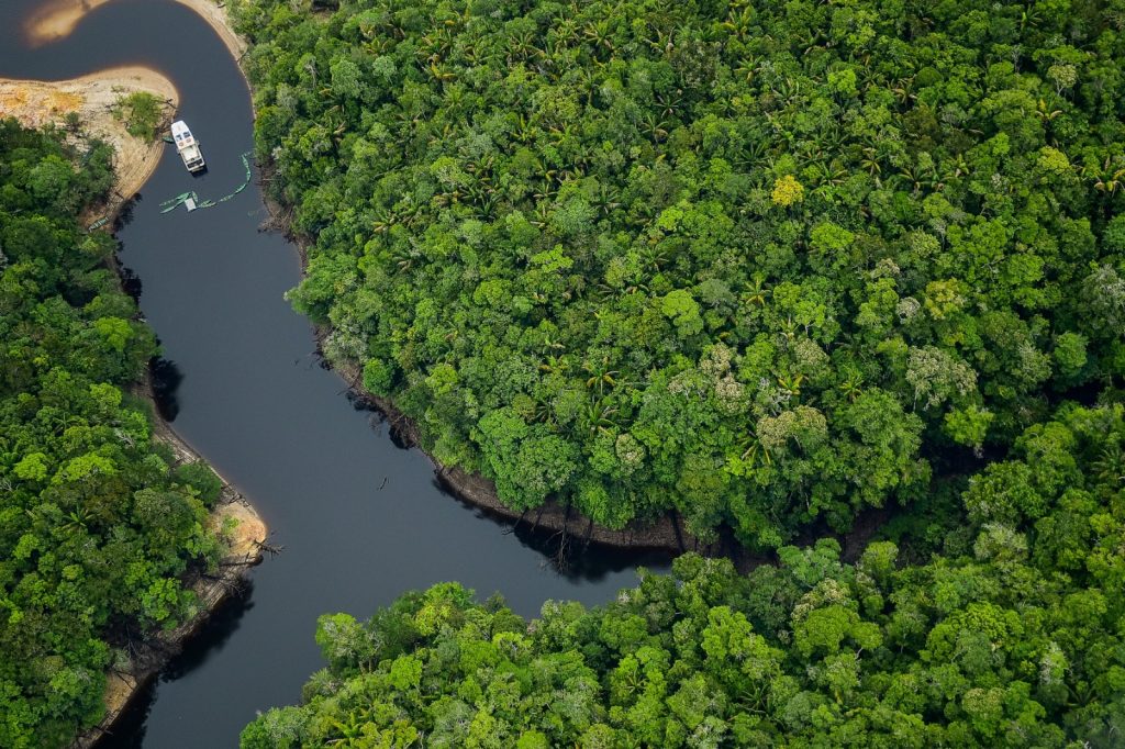 Vista aérea mostra um trecho sinuoso de rio cercado por densa floresta amazônica de copas verdes, com uma pequena embarcação navegando pelas águas escuras.