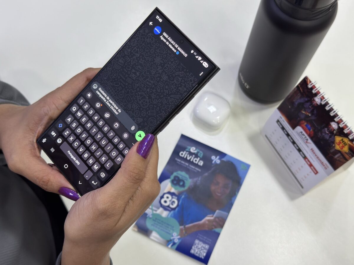 Pessoa usando o WhatsApp no celular para acessar o atendimento da Águas de Manaus, com panfleto da campanha Zera Dívida sobre a mesa.