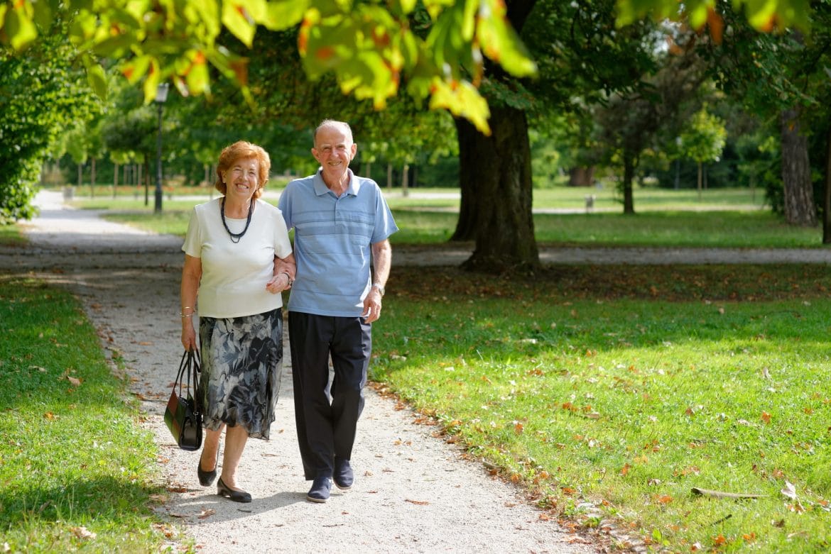 Casal de idosos caminhando em um parque durante o dia, em área arborizada.