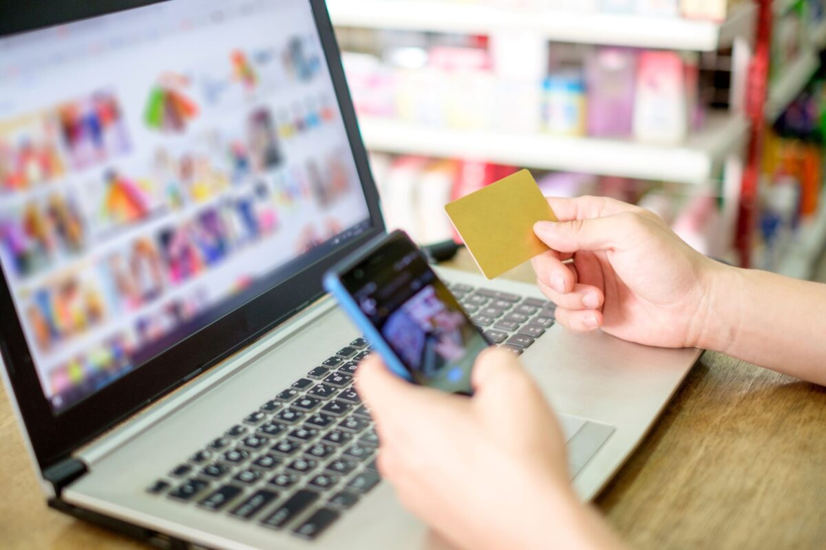 Pessoa utiliza notebook e smartphone para comprar online com cartão em mãos.