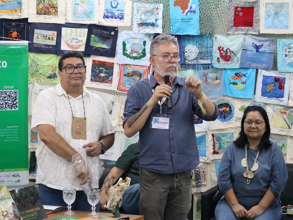 Três participantes falam ao público durante lançamento de livros do Inpa na COP30, diante de painel com trabalhos têxteis.