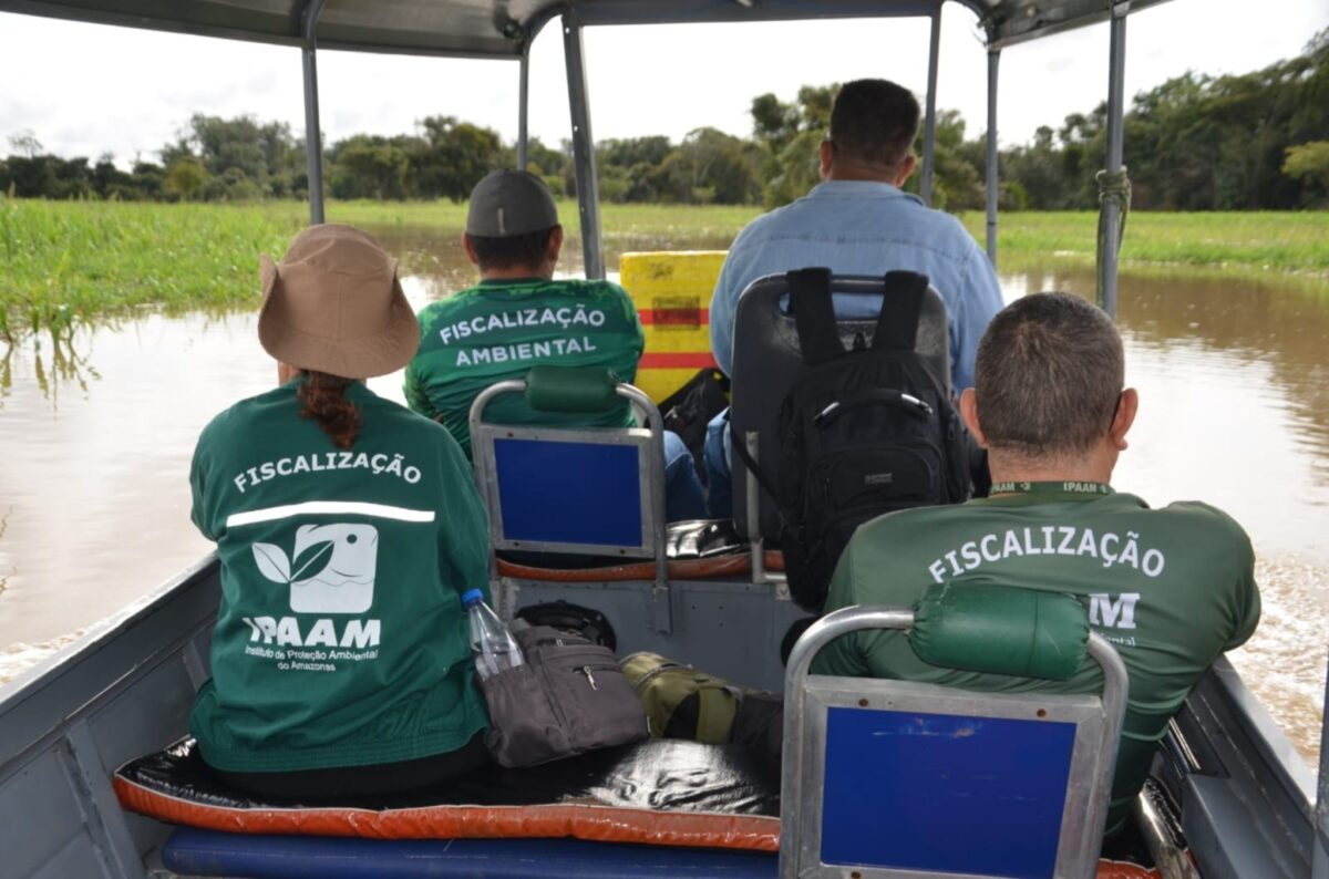 Agentes do Ipaam em barco durante fiscalização ambiental em rio do Amazonas, com área de várzea ao fundo.
