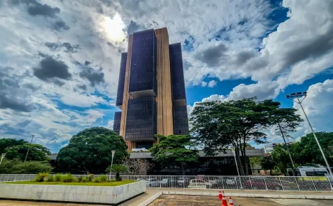 Fachada da sede do Banco Central em Brasília com céu nublado e árvores ao redor.