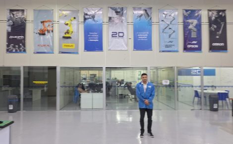 David Cunha, fundador da 2D Innovation, posa em frente ao setor administrativo da empresa, com banners de fabricantes de robôs industriais expostos acima.