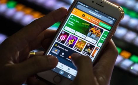 Pessoa utiliza smartphone para acessar aplicativo de apostas esportivas em ambiente com telas iluminadas ao fundo