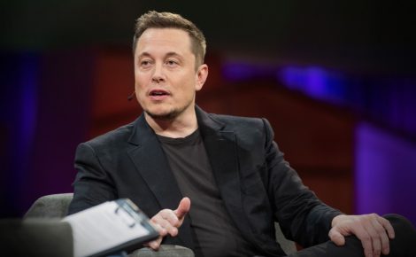 Elon Musk fala durante conferência sobre tecnologia, vestindo terno escuro e gesticulando no palco.