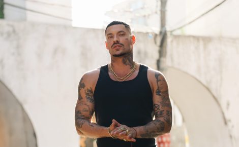 O rapper Filipe Ret posa ao ar livre, vestindo regata preta e calça camuflada, com expressão séria e tatuagens à mostra.
