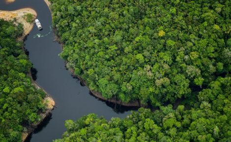 Vista aérea mostra um trecho sinuoso de rio cercado por densa floresta amazônica de copas verdes, com uma pequena embarcação navegando pelas águas escuras.