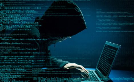 Pessoa encapuzada digitando em um laptop com códigos de programação e números binários em sobreposição, simbolizando cibercrimes e roubo de dados.