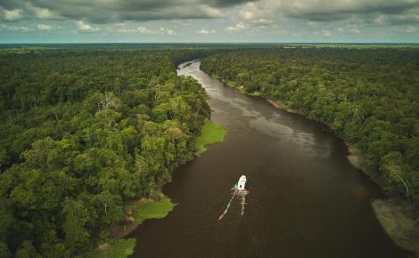 Vista aérea de um barco navegando por um rio sinuoso cercado por floresta densa na Amazônia sob céu nublado.