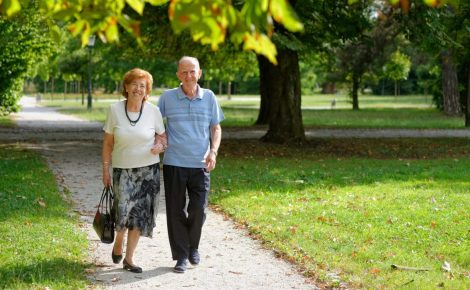 Casal de idosos caminhando em um parque durante o dia, em área arborizada.