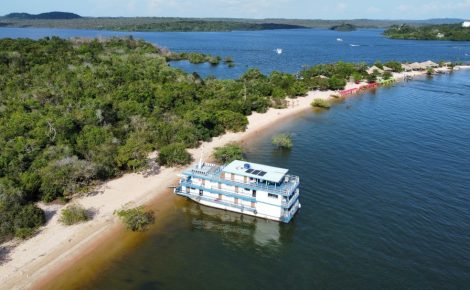Vista aérea de um barco-laboratório atracado em uma praia amazônica cercada por vegetação e rios, durante preparação da Caravana Iaraçu rumo à COP30 em Belém.