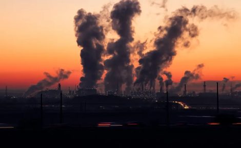 Chaminés industriais liberando grande quantidade de fumaça ao entardecer, com o céu alaranjado ao fundo.