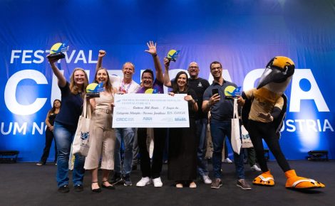 Grupo de vencedores do Festival Curicaca 2025 celebra no palco com troféus e cheque simbólico de R$ 15 mil, ao lado do mascote da competição.