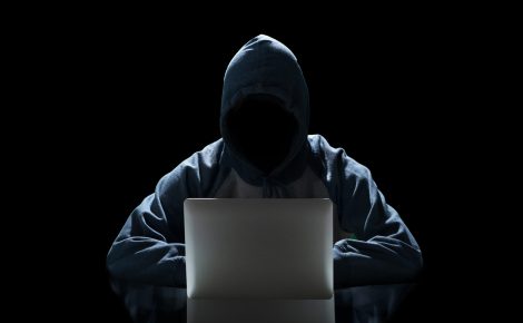 Figura anônima e sombreada de um hacker vestindo um capuz, sentado em frente a um laptop com a tela brilhando, em um cenário escuro, representando a ameaça de ataques cibernéticos