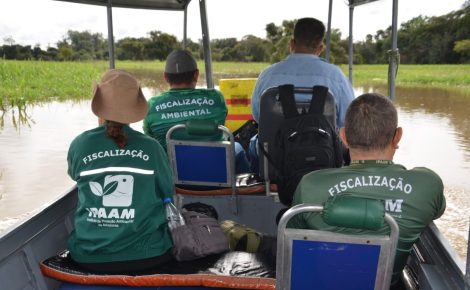 Agentes do Ipaam em barco durante fiscalização ambiental em rio do Amazonas, com área de várzea ao fundo.