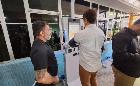 Homem realiza medição de pressão arterial em equipamento K-Doctor enquanto técnico da FPF Tech acompanha o processo em demonstração de triagem médica digital.