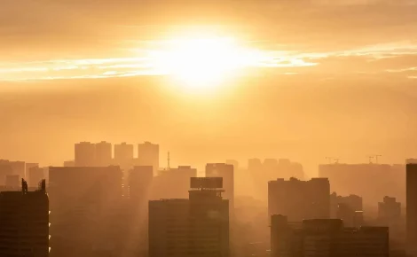 Sol forte iluminando prédios de uma grande cidade sob calor extremo e atmosfera amarelada.