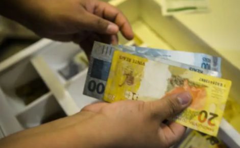 Pessoa contando cédulas de real, com notas de R$ 20, R$ 50 e R$ 100, em ambiente interno.