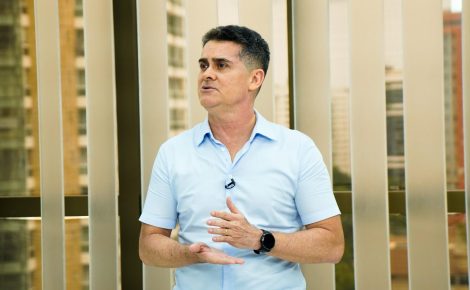 Prefeito David Almeida, de camisa azul-clara, fala em ambiente interno com grandes janelas ao fundo, gesticulando com as mãos.