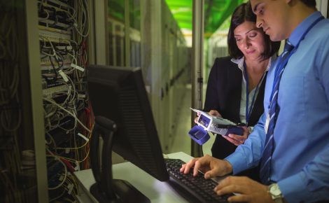 Dois profissionais de TI trabalhando juntos em um computador, com equipamentos eletrônicos e cabos ao fundo, simbolizando a inovação e colaboração no setor.
